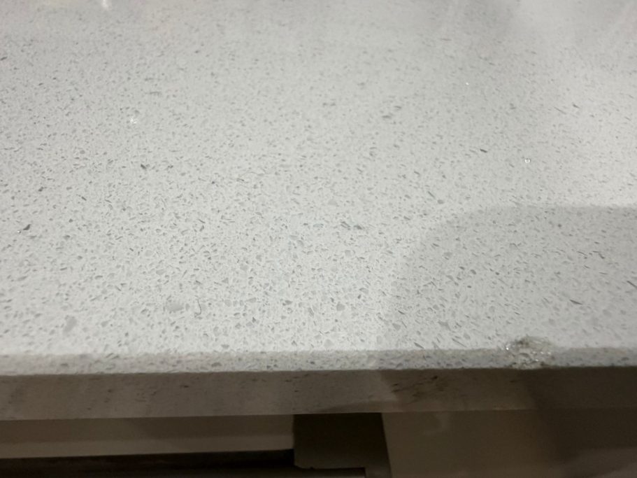 Before repair: quartz edge and surface damage Photo of white quartz worktop showing large chip out of the edge and the surface, damage caused by a plate