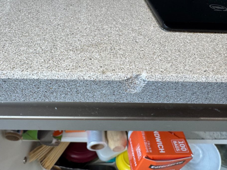 Before repair: chipped grey quartz edge A grey quartz worktop has a significant chip on the edge, and part of the surface is damaged. The damage to the quartz was caused by a plate